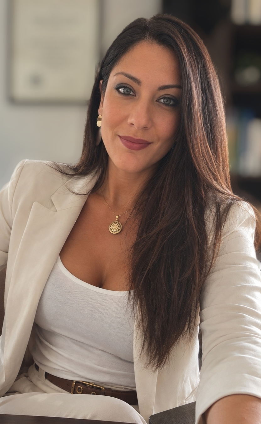 Donna sorridente con capelli lunghi scuri, blazer bianco e collana dorata, seduta in ambiente luminoso.